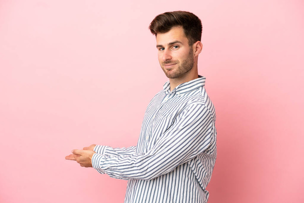 Junger kaukasischer gutaussehender Mann isoliert auf rosa Hintergrund, der die Hände zur Seite ausstreckt, um zum Kommen einzuladen - Foto, Bild