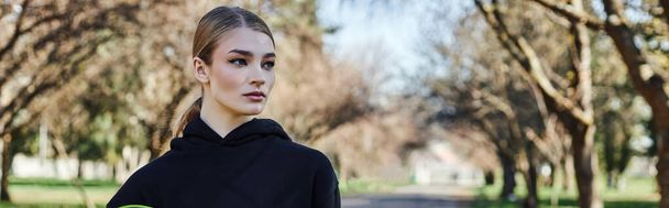 blonde and fit young woman in black sporty hoodie looking at camera after workout in park, banner - Photo, Image