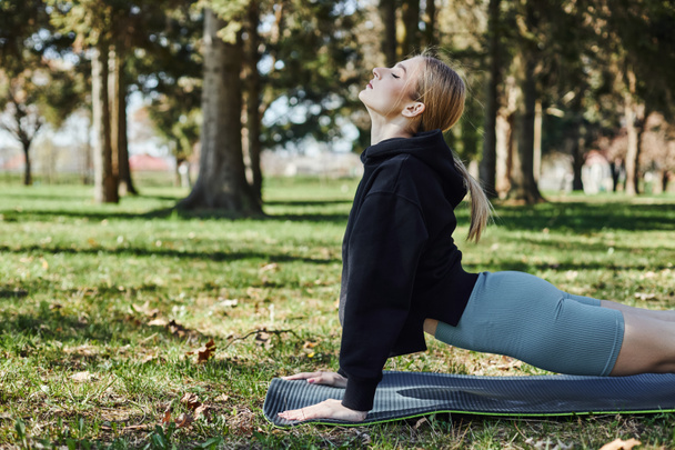 подходит молодая женщина с светлыми волосами и спортивной одеждой медитирует и тянется назад на коврик для йоги в парке - Фото, изображение