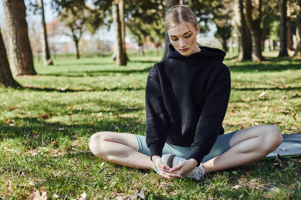 fitte junge Frau mit blonden Haaren und Sportbekleidung sitzt auf Sportmatte und meditiert im Park - Foto, Bild