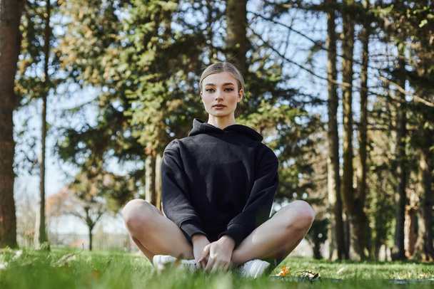 pretty and fit woman with blonde hair and sportswear sitting on mat while meditating in park - Foto, afbeelding
