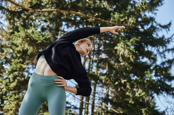 mujer joven y deportiva con cabello rubio y ropa deportiva estirando su cuerpo mientras hace ejercicio en el parque - Foto, Imagen