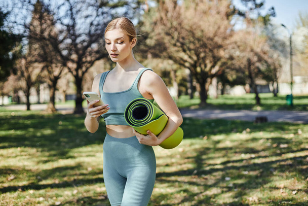 pretty sportswoman in active wear holding smartphone and fitness mat while standing in park - Photo, Image