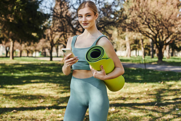 femme sportive heureuse en tenue active tenant smartphone et tapis de fitness tout en se tenant debout dans le parc - Photo, image