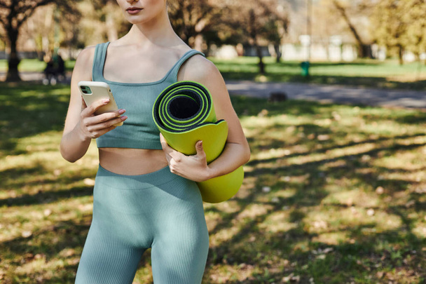 vista recortada de la deportista en desgaste activo celebración de teléfono inteligente y alfombra de fitness en el parque verde - Foto, Imagen