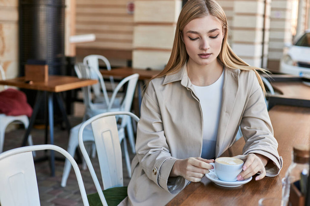 blondýny mladá žena ve stylové a béžové trenč kabát při pohledu na její cappuccino v kavárně, šálek kávy - Fotografie, Obrázek