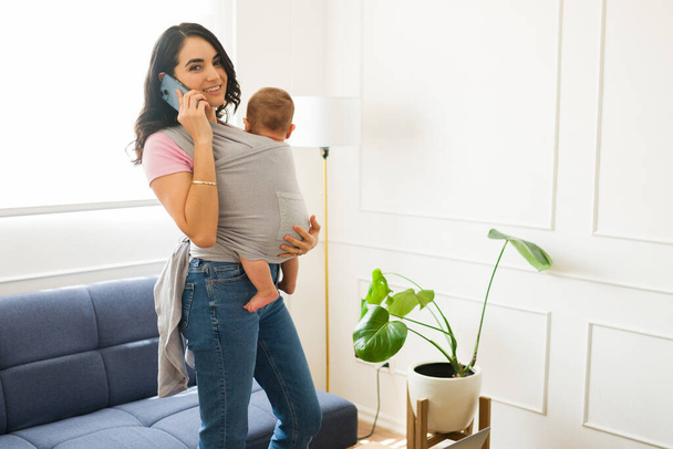 Werken gelukkige moeder ouderschap en het verzorgen van haar pasgeboren baby dragen van haar kind met een baby sling praten aan de telefoon en glimlachen - Foto, afbeelding