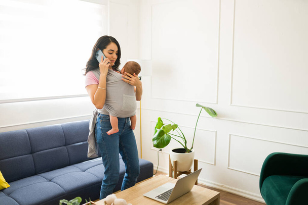 Professional caucasian woman and mom talking on the phone with a customer and working from home carrying her child with a baby sling - Photo, Image