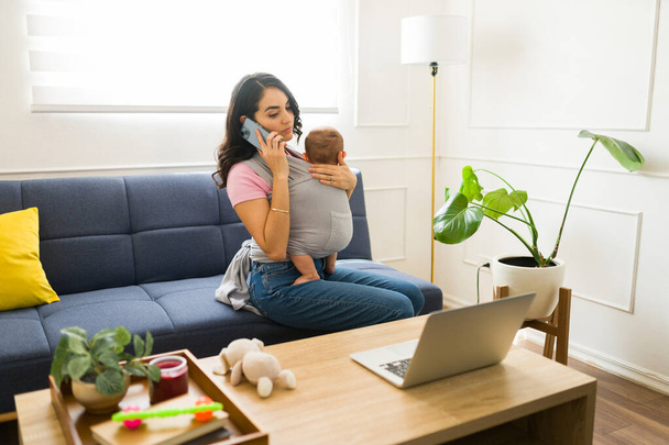 Çekici bir anne çocuğunu kucağında bebek askısı ile evde dizüstü bilgisayarla çalışırken telefonla konuşuyor. - Fotoğraf, Görsel