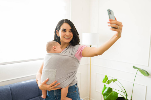 Atractiva mamá feliz tomando una selfie con un teléfono inteligente mientras está criando y llevando a su bebé usando un cabestrillo para bebés - Foto, imagen