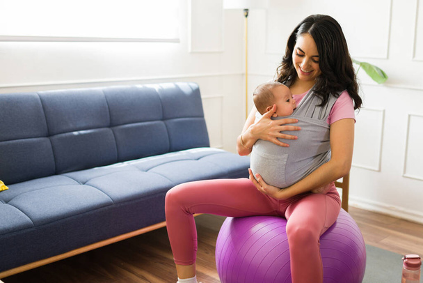 Attraktive, glückliche Mutter trägt ihr Kind in einer Babytrage beim Pilates-Training zu Hause mit einem Stabilitätsball - Foto, Bild
