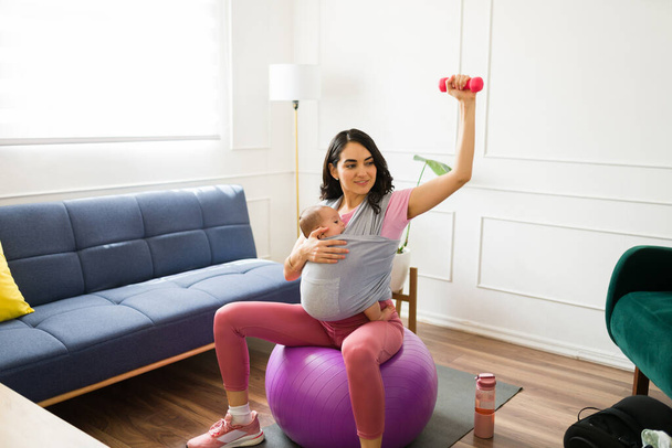 Aktív anya emelő súlyzó súlyzók, miközben csinál egy pilates edzés otthon gyakorló gyermekével egy baba hordozó - Fotó, kép