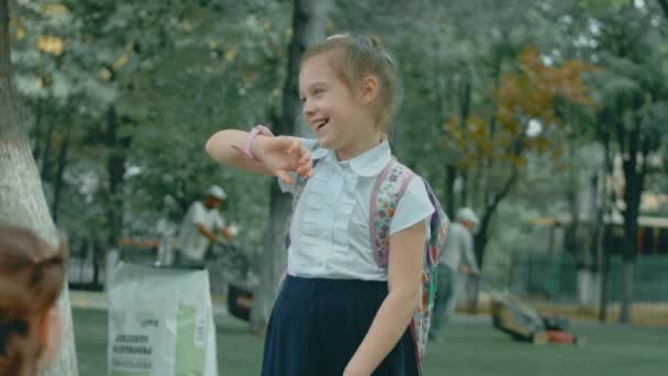 Hermosa chica de la escuela caucásica usando smartwatch al aire libre en el parque escolar después de terminar las clases. Feliz niño hablando con sus padres en una videollamada en el teléfono inteligente. Reloj de pulsera inteligente con rastreador GPS - Imágenes, Vídeo
