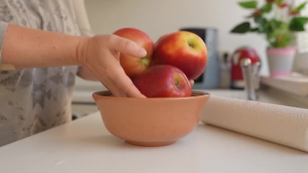 woman puts ripe washed large red apples in ceramic bowl, in home kitchen, preparing for cooking - Footage, Video