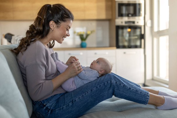 Jeune mère assise sur le canapé à la maison et tenant bébé souriant mignon sur les cuisses et jouer. L'amour maternel, le soin et la joie concept - Photo, image