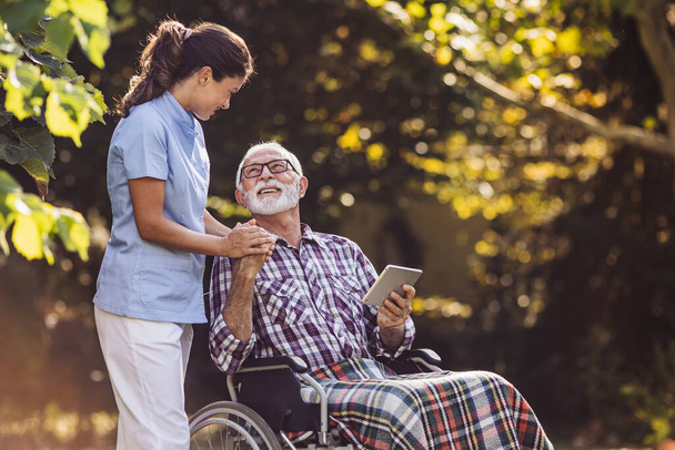 Mladá žena pečovatel pomáhá starší muž na invalidním vozíku s digitálním tabletem a drží jeho ruku v parku - Fotografie, Obrázek