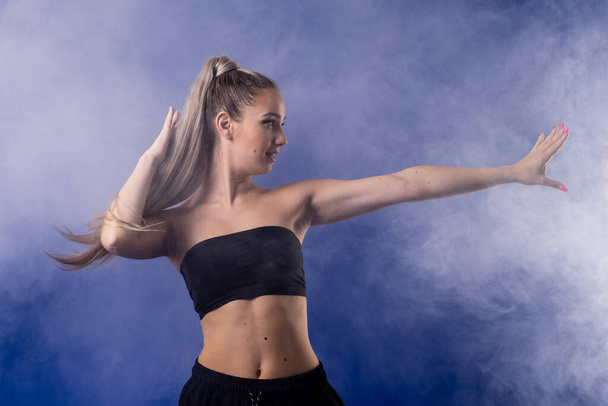 Beautiful blond girl in black outfit dancing zumba against smoke fog and blue background. - Φωτογραφία, εικόνα