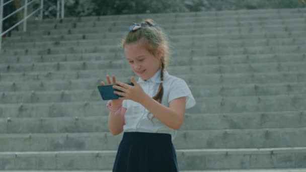 Smiling cute school girl playing with smartphone. Cheerful 8 y.o. girl touching mobile phone with fingers, looking cartoons and having fun with educational apps. Child in park outdoors - Footage, Video
