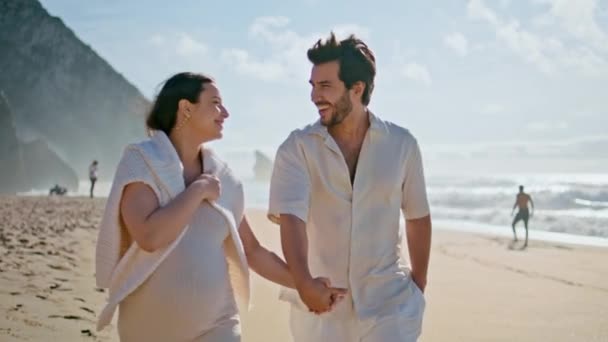 Щаслива вагітна сім'я, що йде красивим літнім морським берегом, тримаючи руки крупним планом. Весела любляча пара сміється, розмовляючи разом на пляжній прогулянці. Безтурботні майбутні батьки насолоджуються відпочинком на морі - Кадри, відео