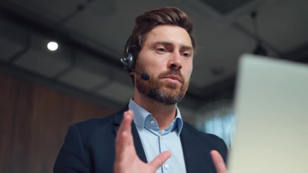 Low angle view of serious bearded man in stylish suit wearing wireless headset sitting at desk with modern laptop and talking during video chat. Concept of business, technology and communication. - Footage, Video