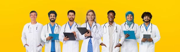 Happy adult, old diverse doctors professional in white coats with tablets, isolated on yellow studio background, panorama. Work in clinic, help online, medicine and health care - Fotografie, Obrázek