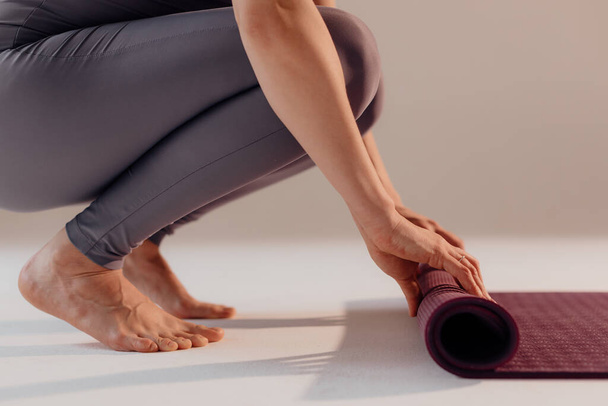 Een jonge vrouw draait een mat voor of na een yogales in de studio. Close-up benen van een vrouw doen yoga terwijl zitten op een lichte achtergrond. - Foto, afbeelding