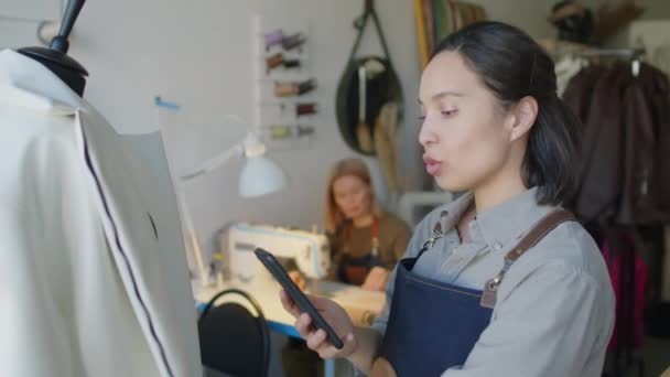 Μέτριο κοντινό πλάνο της νεαρής γυναίκας ράφτη με smartphone μιλάμε για τον πελάτη σε βίντεο, δείχνει ράμματα και το σχεδιασμό της ημιτελές δερμάτινο σακάκι, και συνάδελφος που εργάζονται σε ραπτομηχανή στο παρασκήνιο - Πλάνα, βίντεο