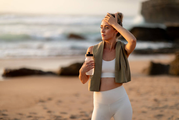 Young tired woman athlete in sportswear holding bottle of water, resting after workout on sea beach, free space. Break, active lifestyle, health care, sport and aqua balance - Photo, Image