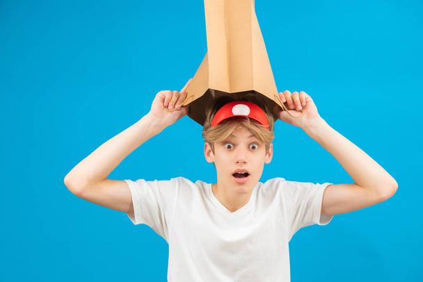 Shocked seller teen boy with paper bag over head. Teenager holding paper bag over the head posing in studio. - Zdjęcie, obraz