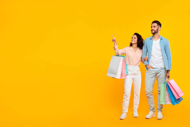 Sezónní slevy. Happy manželé stojící s nákupními taškami a ukazuje stranou na volné místo, pózování na žlutém pozadí, evropský pár těší nakupující obchody, kopírovací prostor - Fotografie, Obrázek