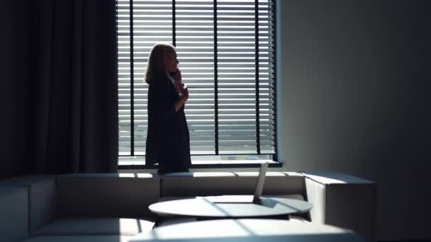 Sivunäkymä vakava valkoihoinen nainen työpuku seisoo lähellä toimistoa ikkuna ja puhuu moderni älypuhelin. Luotettava nainen elehtii emotionaalisesti aikana intensiivistä keskustelua kumppanin kanssa. - Materiaali, video