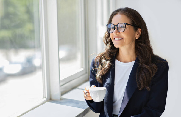 Επιτυχημένη χαρούμενη ελκυστική ώριμη επιχειρηματίας με επίσημο ένδυμα φορώντας γυαλιά ηλίου που στέκεται δίπλα στο παράθυρο με κούπα στο χέρι της, πίνοντας καφέ στο γραφείο, απολαμβάνοντας θέα, αντιγραφή χώρου - Φωτογραφία, εικόνα
