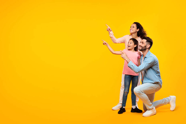 Захоплена сім'я з трьох, що вказують на вільний простір, позуючи над жовтим студійним фоном, повною довжиною, банером. Батьки і дитина дівчина показують і рекламують ваш текст або дизайн - Фото, зображення