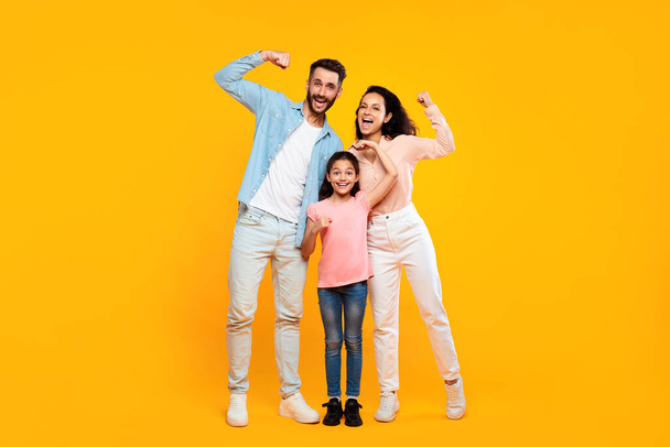 Fuerza familiar. feliz europeo padres y hija mostrando bíceps músculos y sonriendo a la cámara, posando juntos de pie sobre amarillo estudio fondo, longitud completa - Foto, Imagen