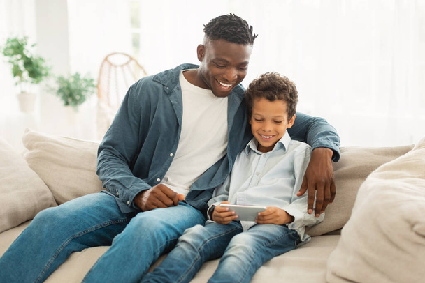 Happy Black Daddy And Little Son Relaxing With Smartphone At Home, Sitting On Couch Indoor and Websurfing Via Phone Gadget, Watching Videos Online On Weekend. Technology And Family Lifestyle - Photo, Image