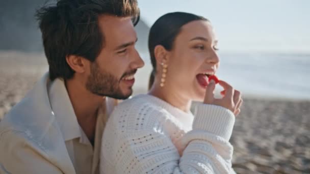Szczęśliwa kobieta jedząca jagodowe przytulanie męża na piaszczystej plaży. Beztroska spokojna para ciesząca się rodzinnym piknikiem na słonecznym brzegu morza patrząc na fale morskie. Przystojny mężczyzna przytula ukochaną kobietę na wybrzeżu - Materiał filmowy, wideo