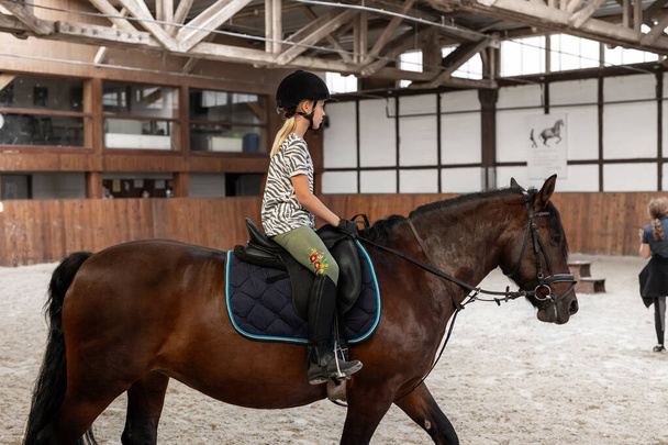 Horse riding school. Little children girls at group training equestrian lessons in indoor ranch horse riding hall. Cute little beginner blond girl kid in helmet sitting on brown horse horseback. - Photo, Image