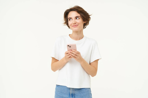 Портрет брюнетки з використанням смартфона, що стоїть з мобільним телефоном, смс, розміщує замовлення, переглядає соціальні медіа по телефону, стоїть на білому тлі. - Фото, зображення