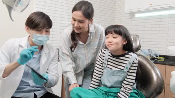 El dentista pediátrico masculino asiático explica la radiografía de los dientes a la niña y a su madre para la higiene dental desde la tableta en la clínica dental, el médico oral profesional de ortodoncia en un hospital de atención médica infantil. - Metraje, vídeo