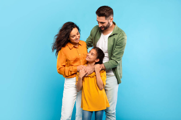 Young parents and little daughter posing hugging and smiling to each other standing together on blue background, studio shot. Happy european family of three - Photo, Image