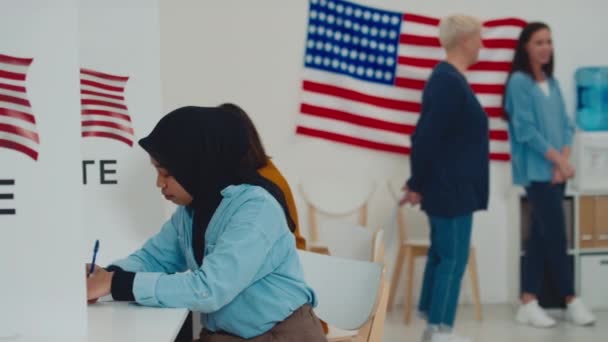Mediana toma de una joven musulmana en hiyab islámico negro sentada en cabina en la estación electoral y llenando su boleta electoral, y señoras irreconocibles charlando en un fondo borroso junto a la bandera de EE.UU. - Imágenes, Vídeo