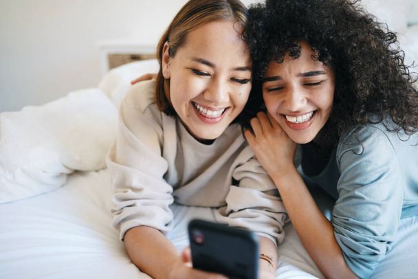 Handy, Entspannung und junges lesbisches Paar im Bett vernetzen sich in sozialen Medien, mobilen Apps oder im Internet. Glückliche, technologische und interrassische lgbtq-Frauen scrollen auf einem Telefon im Schlafzimmer einer modernen Wohnung. - Foto, Bild