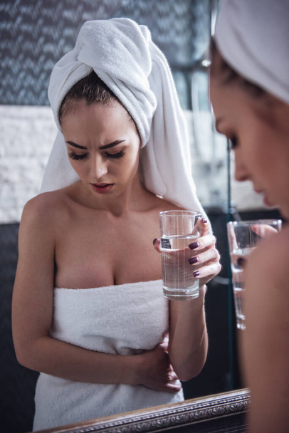 Menina bonita na toalha de banho está sentindo dor, segurando um copo de água e olhando para o espelho no banheiro
 - Foto, Imagem