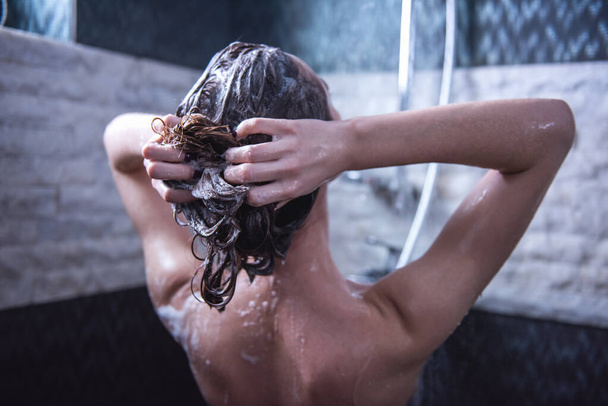 浴室でシャワーを浴びながら彼女の髪を洗う美しい裸の若い女性の背面図 - 写真・画像