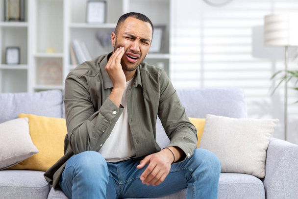 Jeune homme hispanique souffrant de maux de dents assis sur le canapé à la maison et tenant la main à la joue. - Photo, image