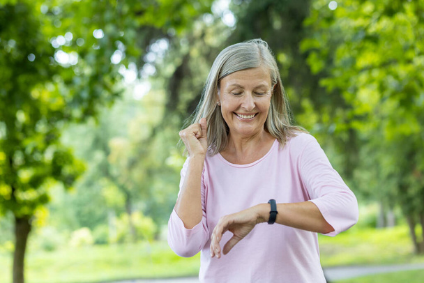 Senior gray-haired beautiful woman is running and doing sports in the park and checking the time on the tracker, happy with the result. He looks at the smart watch on his wrist. - Photo, image