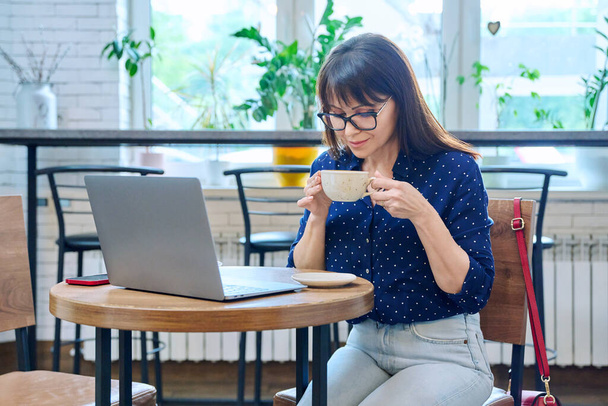 Middle-aged woman drinking coffee looking at laptop screen, sitting at table in cafe, coffee shop. Remote work, freelancing, blogging, chatting, online internet technology, mature people concept - Photo, Image