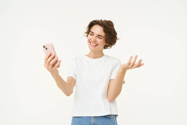 Το Potrait of woman video chats with friend, δείχνοντας κάτι, έχει μια online συνάντηση στο smartphone, στέκεται πάνω από το λευκό φόντο. - Φωτογραφία, εικόνα