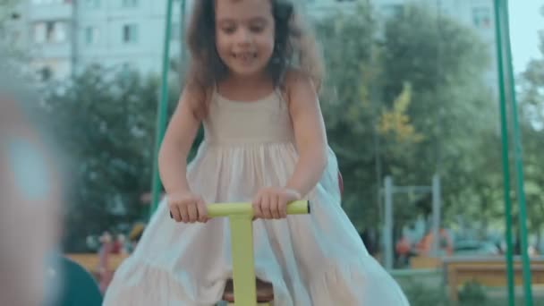 Glückliche kleine kaukasische Mädchen sitzen an einem schönen Sommertag auf einer Metallschaukel auf dem Spielplatz - Filmmaterial, Video