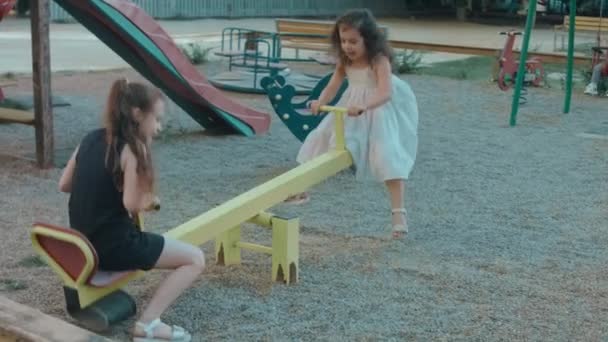 Feliz poco caucásico niñas sentarse en un metal meciéndose en el patio de recreo en un hermoso día de verano - Imágenes, Vídeo